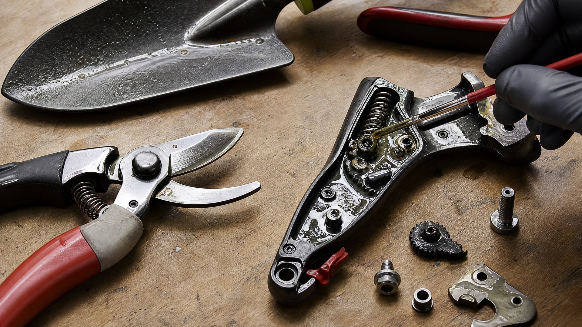 На верстаке лежат садовые ножницы и лопатка, обработанные смазкой, рядом разобранный инструмент.
