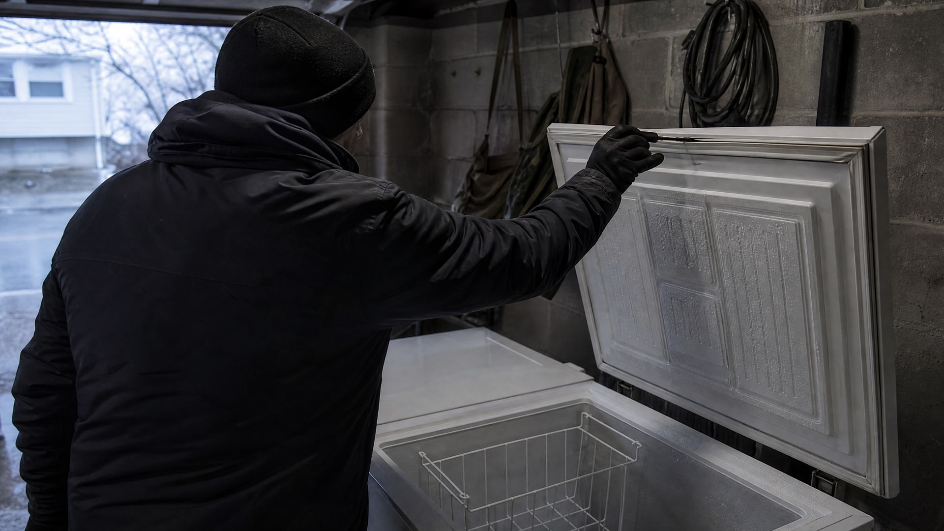 Человек в куртке наносит прозрачную смазку на уплотнитель морозильной камеры в гараже.